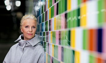 A blonde woman dressed in gray stands next to a colourful wall looking straight at the camera