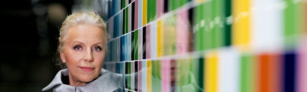 A blonde woman dressed in gray stands next to a colourful wall looking straight at the camera