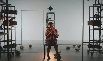 a woman sits in a sparse white room with watermelon halves on the floor around her