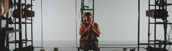 a woman sits in a sparse white room with watermelon halves on the floor around her