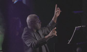 A man sits at a piano conducting with his hands in the air