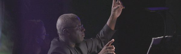 A man sits at a piano conducting with his hands in the air