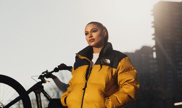 A woman stands in early-morning sunlight looking down into the camera, with her hair slicked back into a low ponytail and her hands in the pockets of her yellow puffa jacket. A bicycle wheel and tower block are visible behind here.