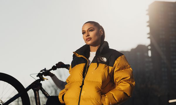 A woman stands in early-morning sunlight looking down into the camera, with her hair slicked back into a low ponytail and her hands in the pockets of her yellow puffa jacket. A bicycle wheel and tower block are visible behind here.