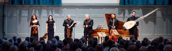 Six musicians in black stand on the stage with their instruments after a concert. The walls and curtains behind them are blue and in front of them a full concert hall of people is applauding.