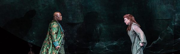 A man and woman, both dressed in elaborate robes, sing to each other on a dark, watery stage, facing each other with intent expressions.
