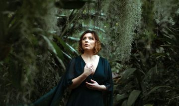 A woman in a flowing green dress stands in a dark forest with moss trailing around her. She is looking off to the side.