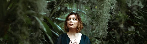 A woman in a flowing green dress stands in a dark forest with moss trailing around her. She is looking off to the side.