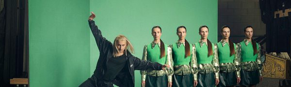 A row of identical women stand emotionless behind a male dancer who appears mid-movement, with his arms outstretched and weight balanced on one leg.
