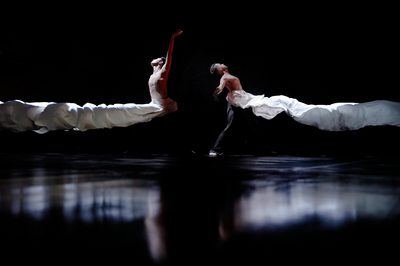 Two men stand back to back on a dark stage, throwing their arms in the air as long streams white cloth billow away from them