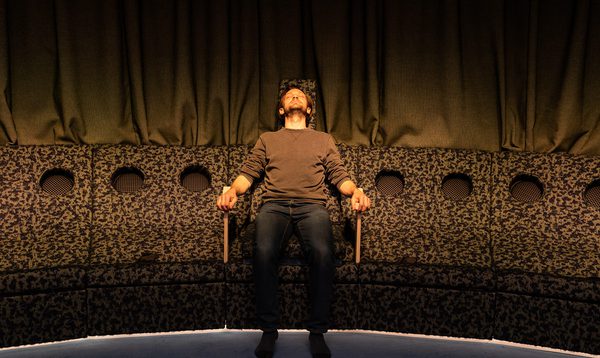 A man sits in the centre of a row of curved seats in an upright position with his arms resting on armrests, gazing up at a bright line shining down onto his face.