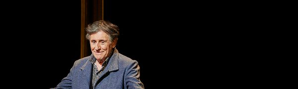 A smiling man stands in the spotlight on a dark stage, wearing a blue coat with his arms outstretched.