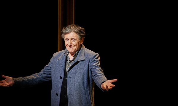 A smiling man stands in the spotlight on a dark stage, wearing a blue coat with his arms outstretched.