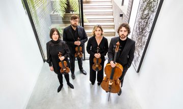 Two men and two women, all dressed in black, stand looking at the camera with serious faces, each hold a string instrument