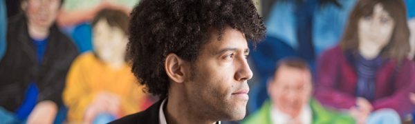 A man with light brown skin and short curly hair sits in the middle of the image wearing a suit and shirt. He looks to the side and behind him is a colourful, blurred out mural of lots of sitting people.