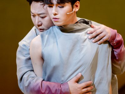 A singer in grey clothing is caught in an embrace. The man that hugs them from behind has red stained forearms.