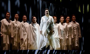 Nine women dressed in white stand solemnly in the shape of a triangle. The woman at the front is standing in a beam of light.