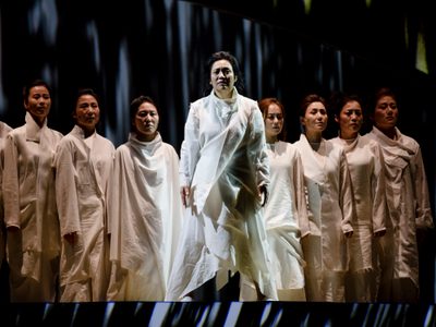 Nine women dressed in white stand solemnly in the shape of a triangle. The woman at the front is standing in a beam of light.