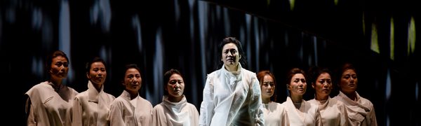 Nine women dressed in white stand solemnly in the shape of a triangle. The woman at the front is standing in a beam of light.