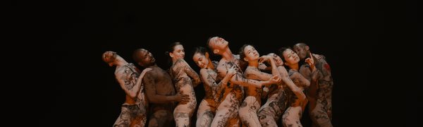 Nine dancers in skin-coloured suits with tattoos on them stand entangled in a line