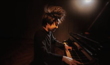 Mao Fujita plays a grand piano furiously, so much so that his hair stands on end. He is bathed in a spot of warm orange light
