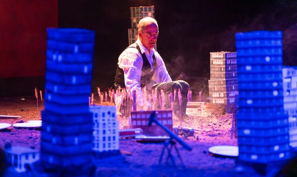 An actor wearing a white shirt and waistcoat sits immersed in dirt. Around him are model city buildings, trucks and oil rigs., tr