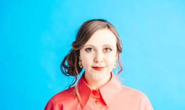 A woman wears a coral button down shirt, in front of a bright blue background. Her hair is tied loosely to the side, and she smiles softly at the camera.