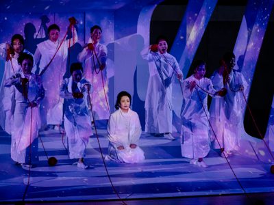 Eight women stand singing defiantly while they are pulling red yarn. In the middle sits a woman on her knees, while images of the universe is projected on them.