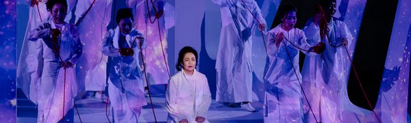 Eight women stand singing defiantly while they are pulling red yarn. In the middle sits a woman on her knees, while images of the universe is projected on them.