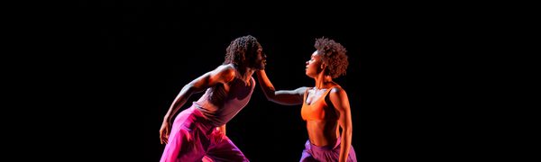 Two dancers bathed in a neon light against a black background lean towards each other as one cups the other's cheek with their palm