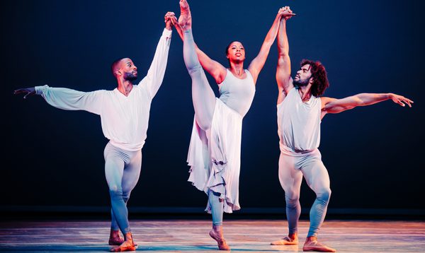 Three dancers perform on stage. One holds one leg high in the air, as she is supported on either side by two male dancers
