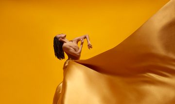 A woman stands against a yellow background with a golden silk dress billowing around her and her head tipped back with dreadlocks hanging down