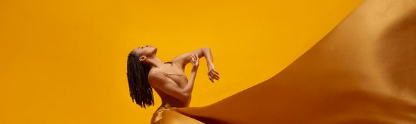 A woman stands against a yellow background with a golden silk dress billowing around her and her head tipped back with dreadlocks hanging down
