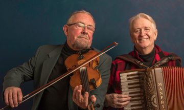 Two men sit side by side, one playing a fiddle with his eyes closed and one playing an accordion and smiling at the camera