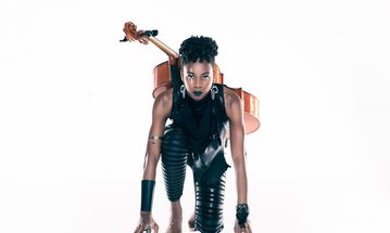 A woman crouches with a cello on her back, shes dressed in black leather and has cuffs on her wrists and bicep. Her makeup is dramatic, with black lipstick and a white line across one eye.