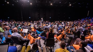 An audience sit on blue and orange beanbags with orchestra players seated on chairs amongst them and a conductor standing in the middle