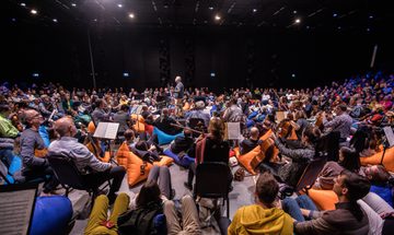 An audience sit on blue and orange beanbags with orchestra players seated on chairs amongst them and a conductor standing in the middle