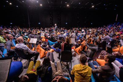 An audience sit on blue and orange beanbags with orchestra players seated on chairs amongst them and a conductor standing in the middle