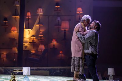 An elderly couple stand in an embrace, illuminated by a wall decorated with table lamps.