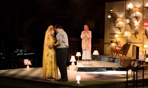 A male and female actor embrace, as they are watched mournfully by an elderly female actor. Behind them is a wall covered in small lamps
