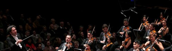 A conductor with curly hair stands on stage conducting an orchestra mid-performance