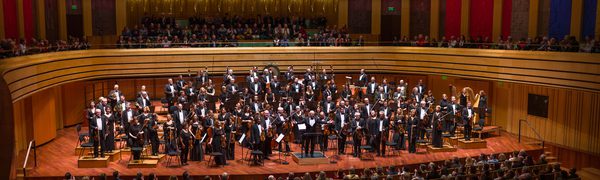 The orchestra pictured standing onstage at the conclusion of a performance, facing their audience and smiling broadly.