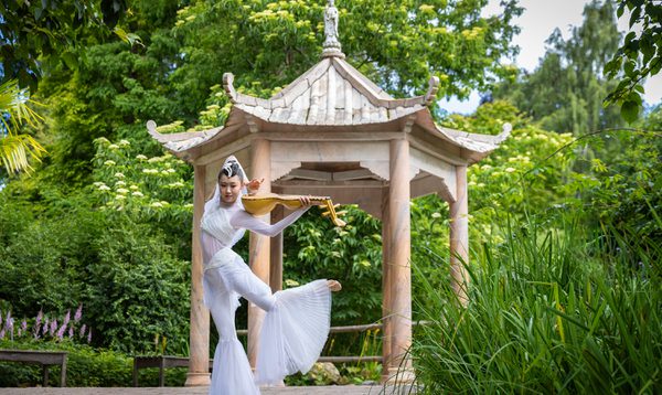 A Chinese dancer holds her instrument and dances in a white costume with flared pants in front of the Chine rotunda at the Edinburgh Botanic Gardens