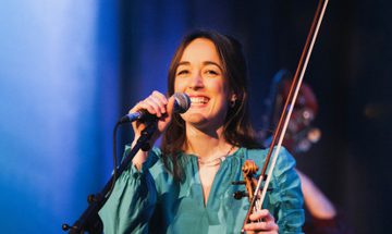 The violinist pictured smiling onstage, holding her violin and bow in one hand and a microphone on a stand in the other.