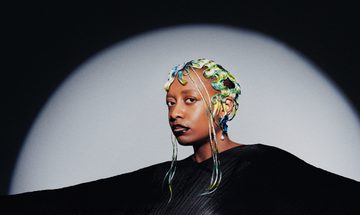 The singer pictured with green and yellow pin curls wearing a black diaphonous gown, illuminated by a spotlight against a black background.