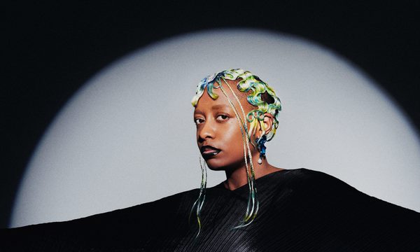The singer pictured with green and yellow pin curls wearing a black diaphonous gown, illuminated by a spotlight against a black background.