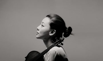 Violinist holds the neck of the violin against her shoulder as she looks ahead, she stands in front of a grey backdrop
