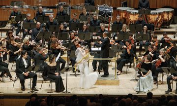 An orchestra on stage with a woman in a white voluminous dress playing a curvacious stringed instrument at the front