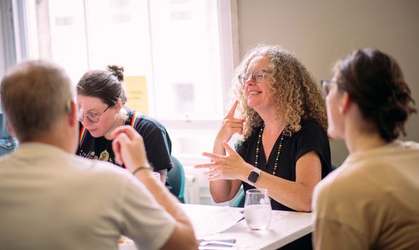 People sit around a table, a woman signs in BSL to another person at the table, she is smiling.