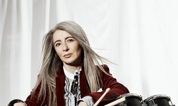 a woman with long grey hair leans against matte black percussion drums. She wears a maroon blazer, and looks at the camera with a serious expression.